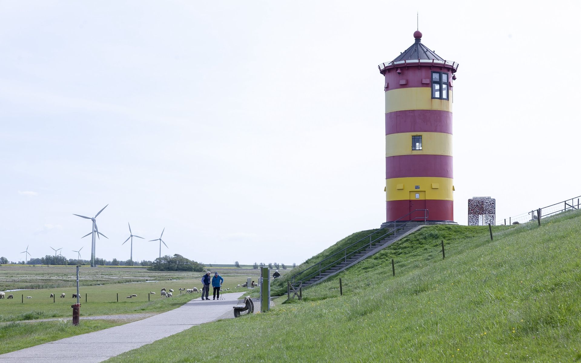Ein rot-gelb gestreifter Leuchtturm steht vor blauem Himmel auf dem grünen Deich.